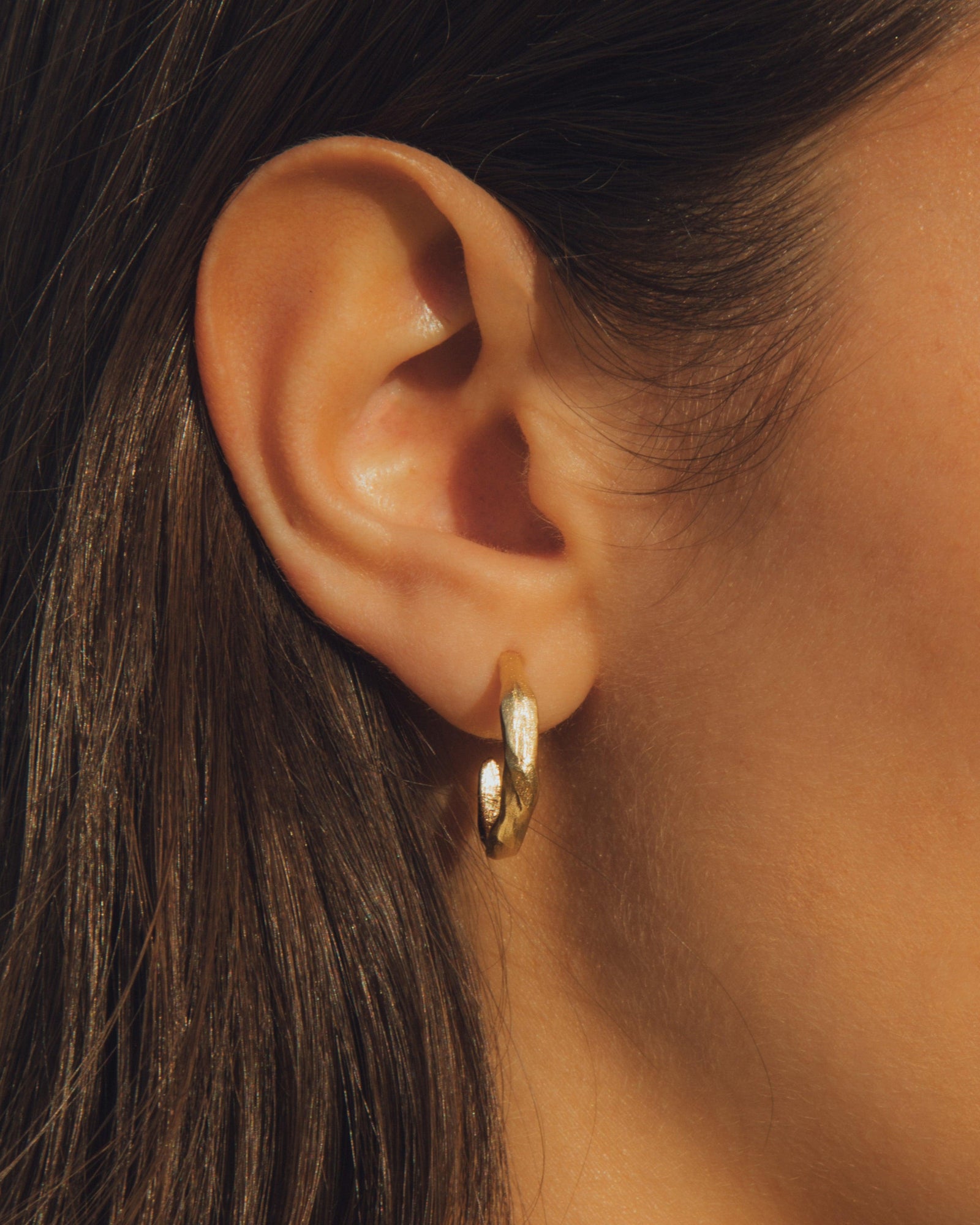 Close-up of a woman's ear wearing a small, irregular gold plated hoop earring.
