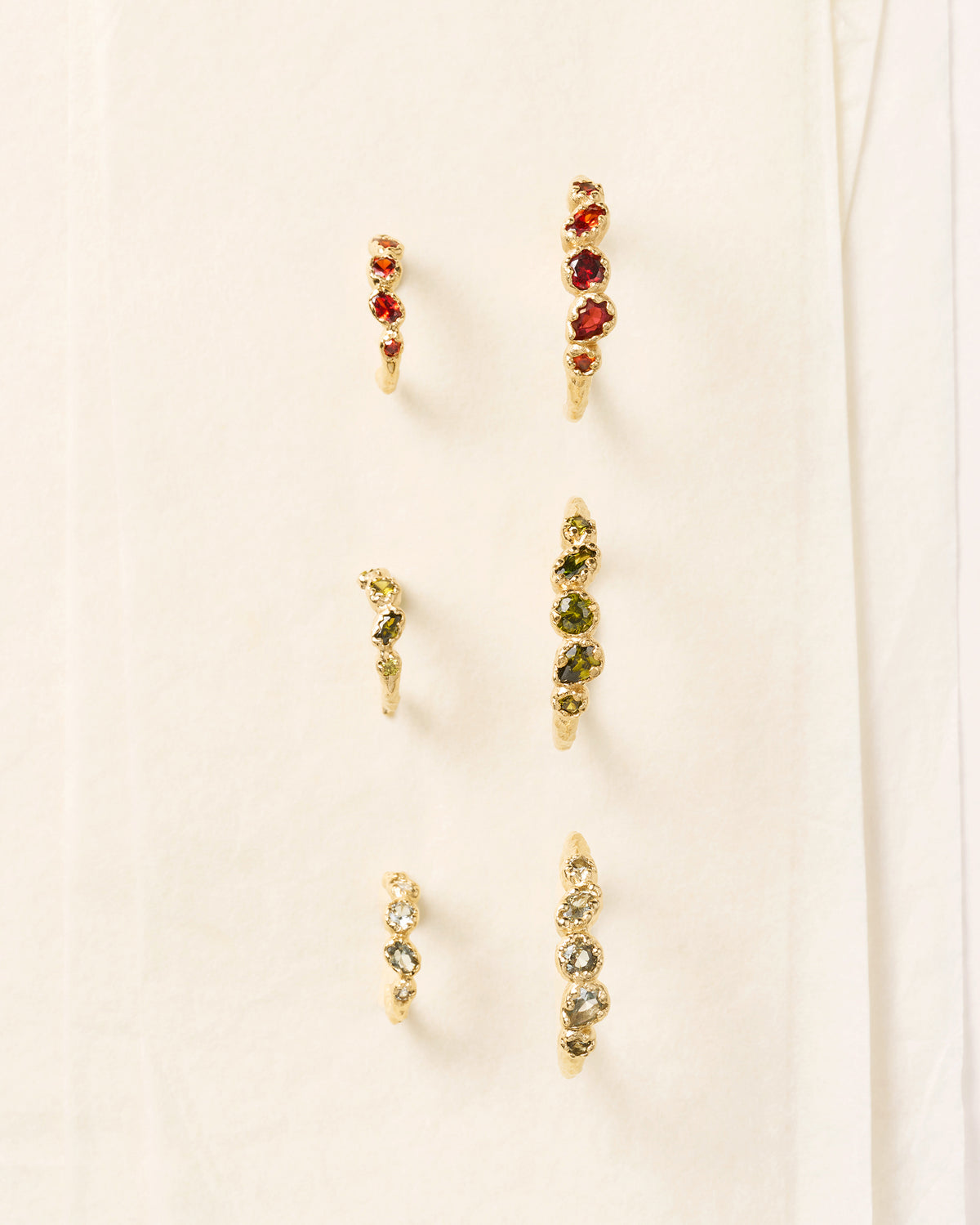 Three pairs of gold hoop earrings with textured bands, featuring red, green, and clear gemstone clusters on a white background.