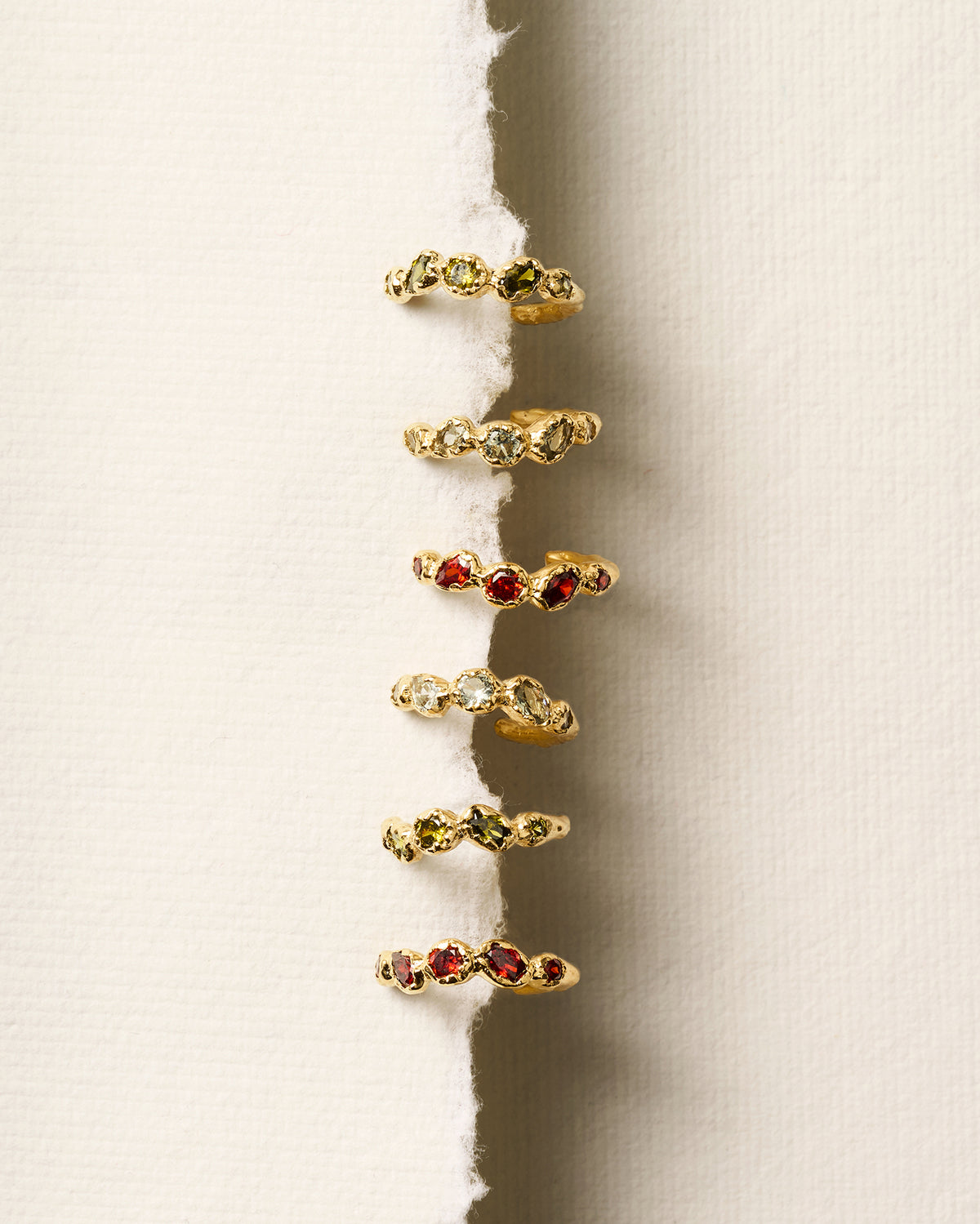 Gold hoop earrings with textured settings and five colorful, hand-set stones arranged vertically on a torn paper edge.