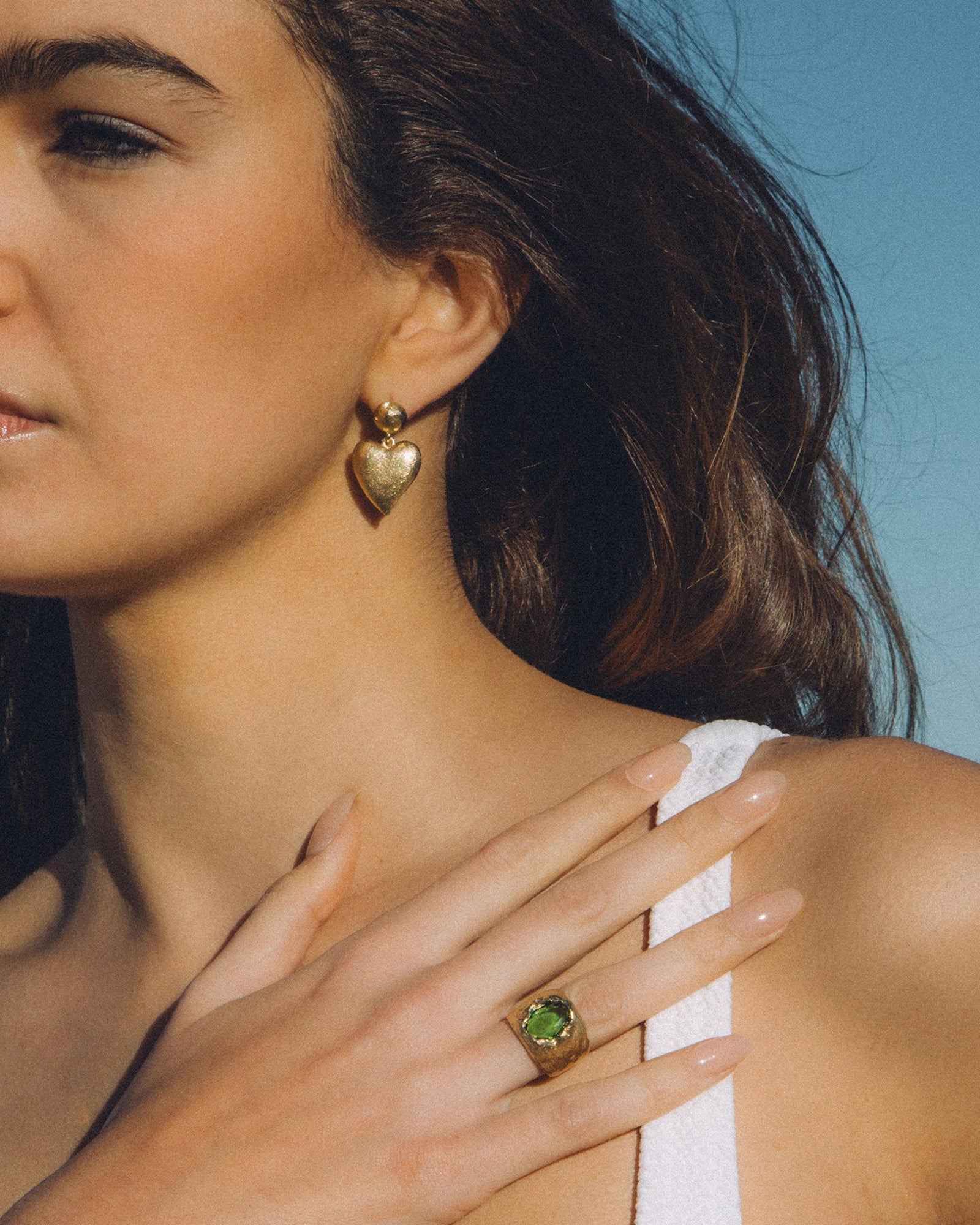 Woman wearing textured gold heart drop earrings and a gold ring with a green gemstone.
