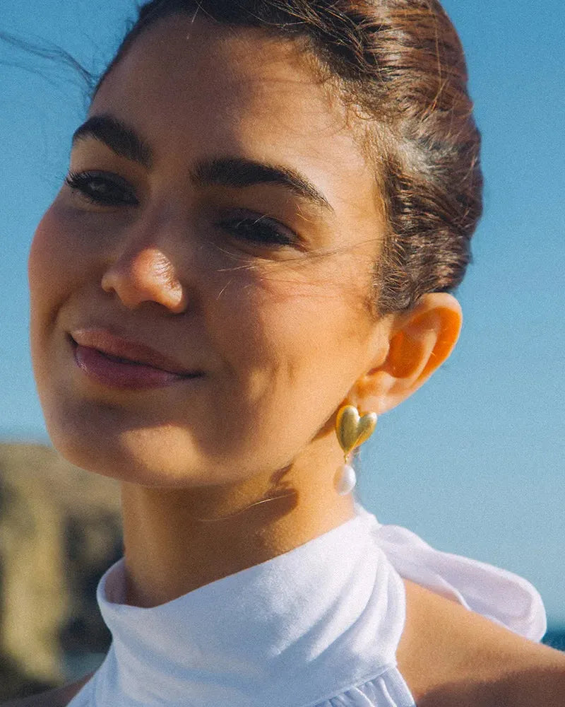Woman wearing a gold heart earring with a dangling white pearl, against a blue sky.