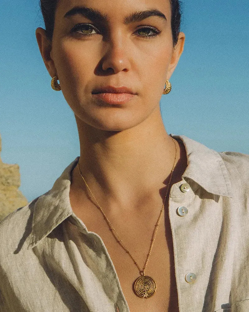 Woman wearing a gold Knossos necklace with a circular labyrinth pendant and matching gold hoop earrings.