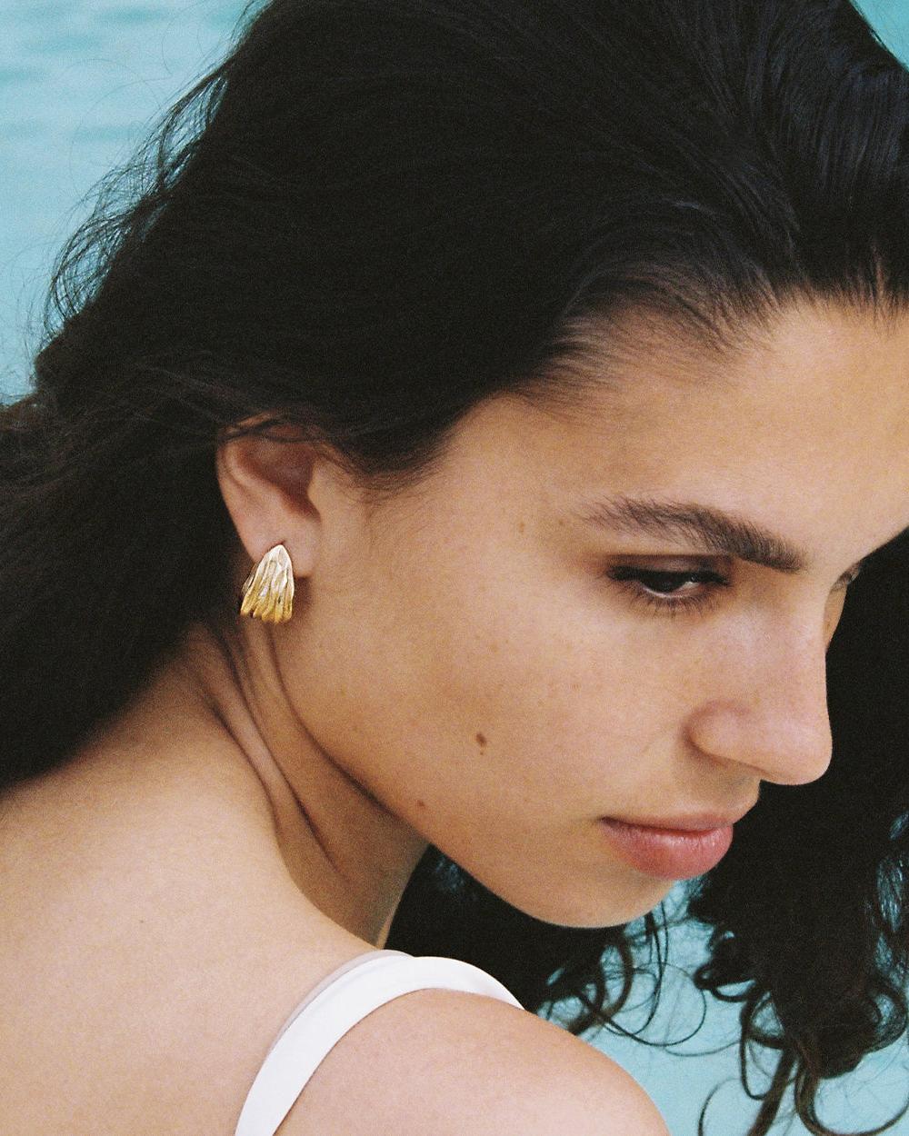 Close-up of a woman wearing a single Lina earring: a small, textured gold stud with an organic, rippled design.