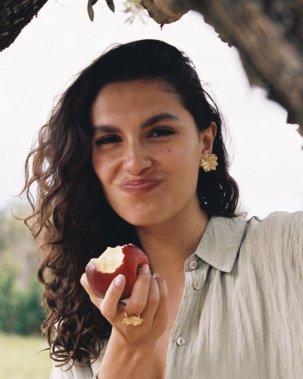Woman smiling, wearing large gold flower-shaped Adonis earrings and a matching ring, holding a bitten red apple.