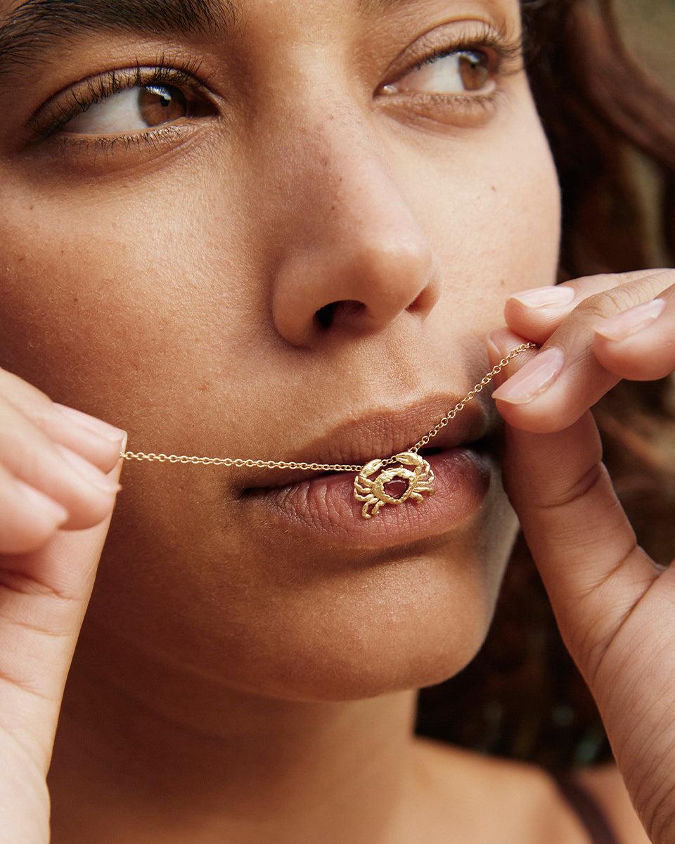 Gold Crabi necklace with a small crab pendant and a delicate red stone heart, held near lips.