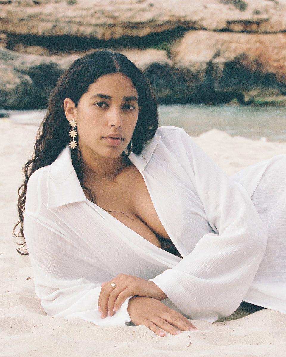 Woman on beach wearing Solara gold sun pendant earrings and a white sheer shirt.