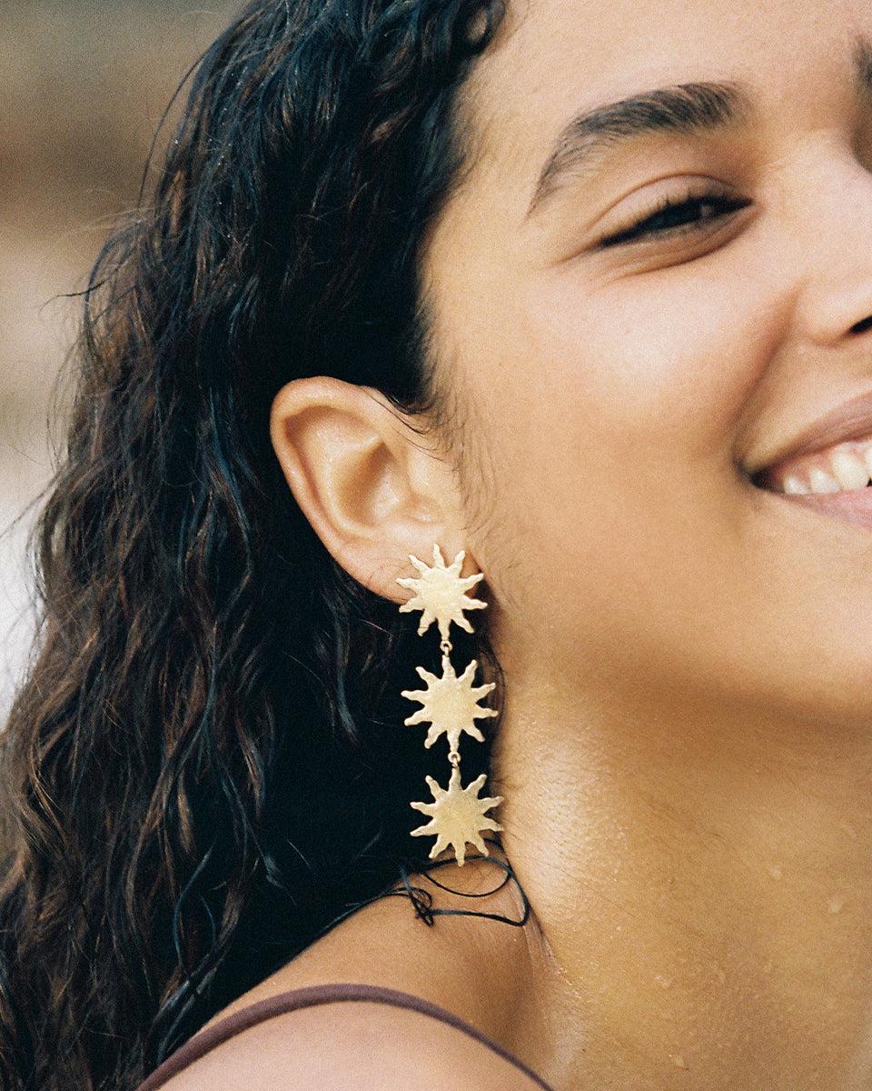 Woman wearing long Solara pendant earrings with three matte gold sunbursts.