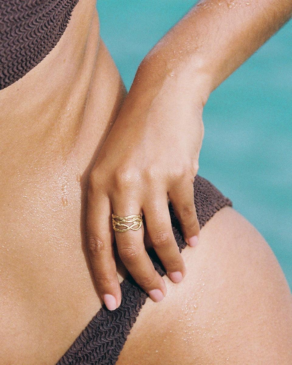 Gold Olea ring with intertwined lines and coral branch design on a hand against a blue background.