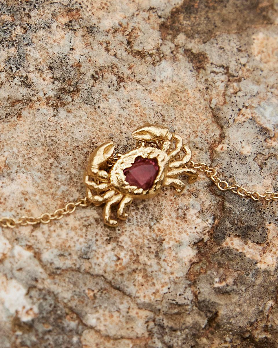 Gold crab pendant necklace with a delicate red heart-shaped stone, resting on a textured rock.