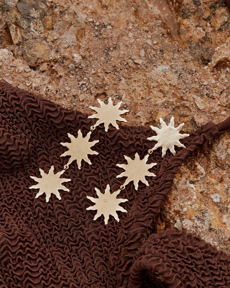 Gold Solara pendant earrings with three suns on a brown textured fabric.