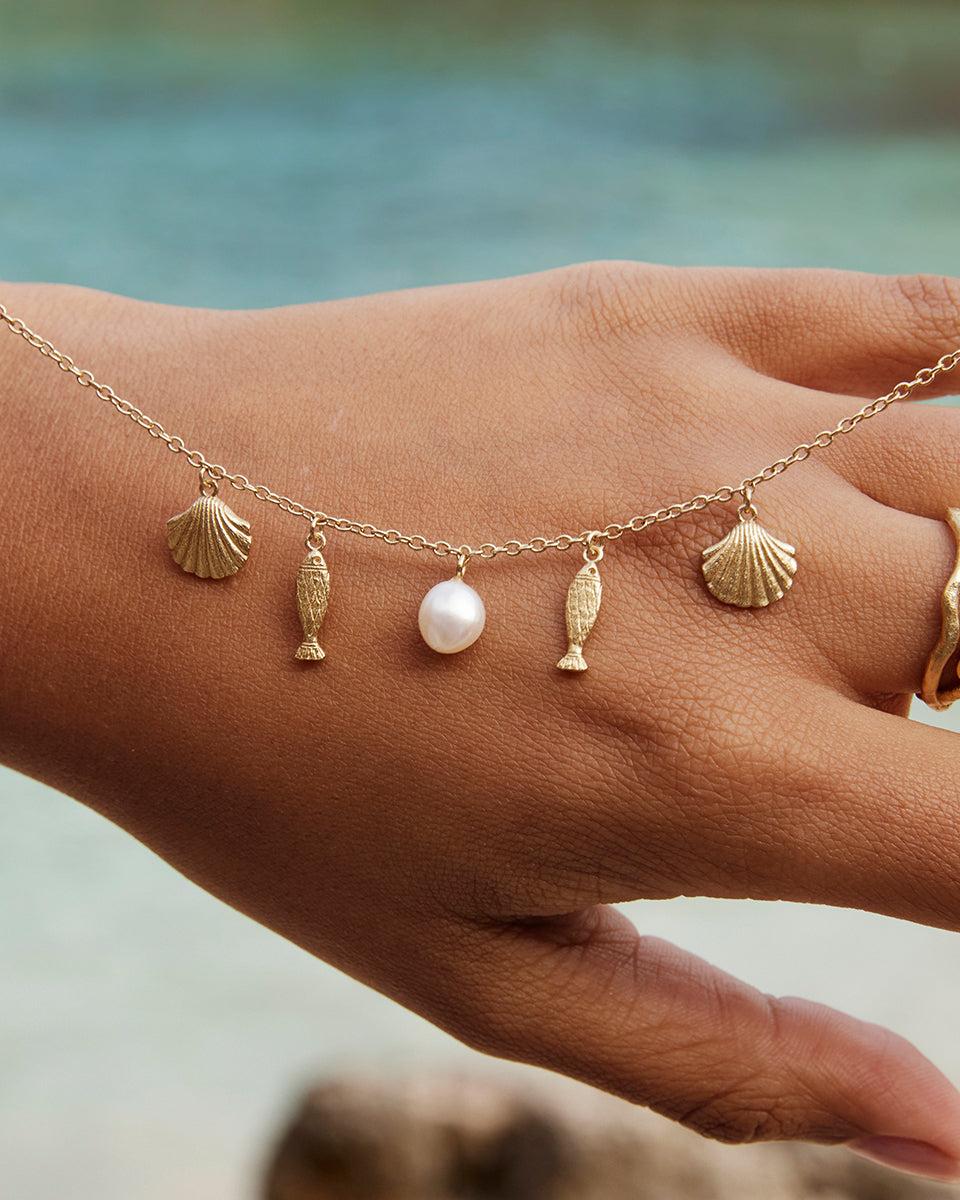 Gold Sardela necklace with seashell, fish, and pearl charms on a hand, ocean background.