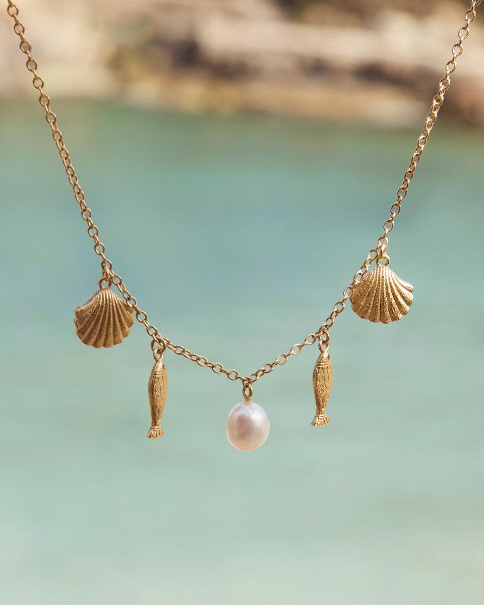 Gold Sardela necklace with seashell, fish, and pearly charms against a blurred blue background.