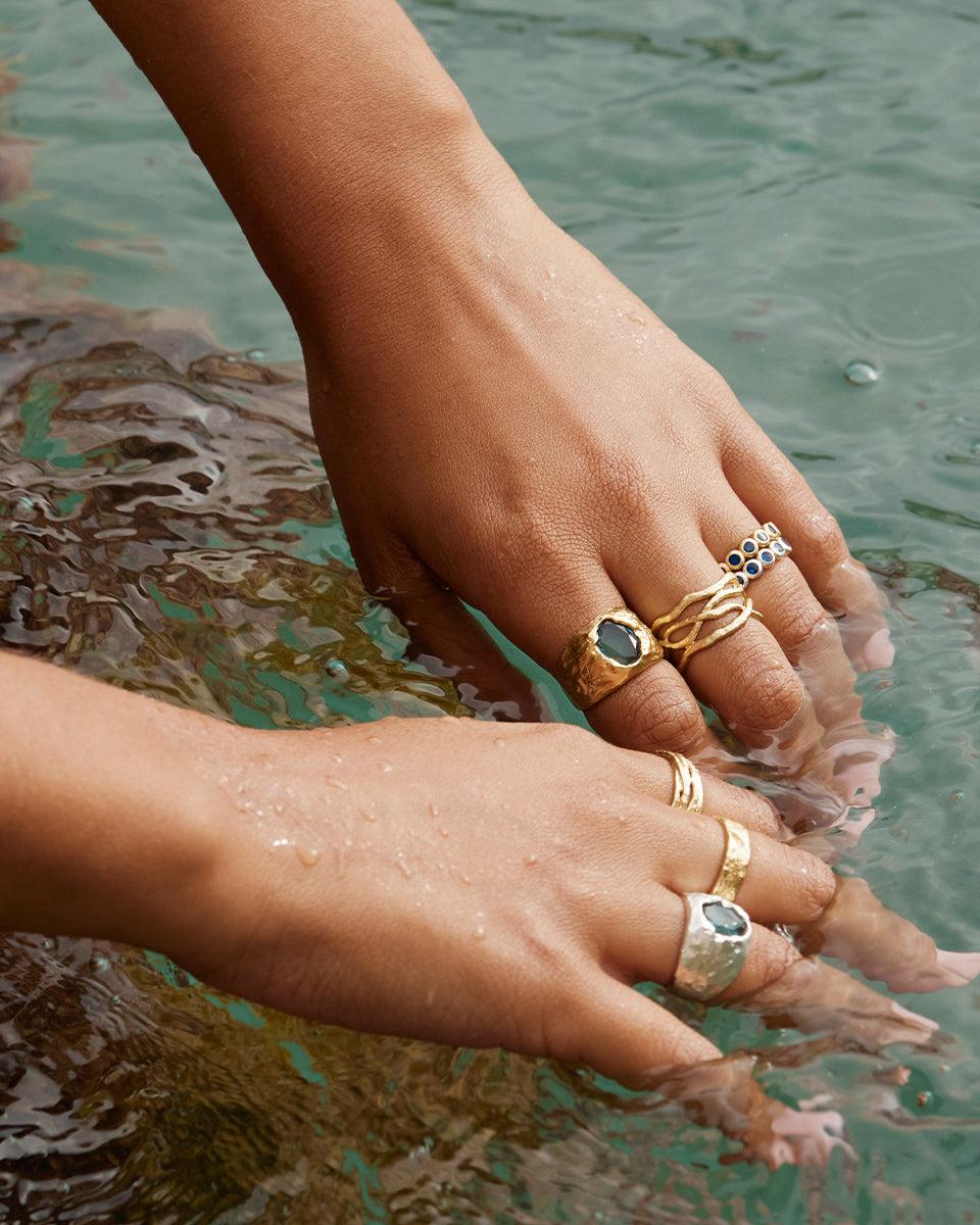 Hands in water wearing Olea ring and other gold rings with blue stones, coral branch, and wave designs.