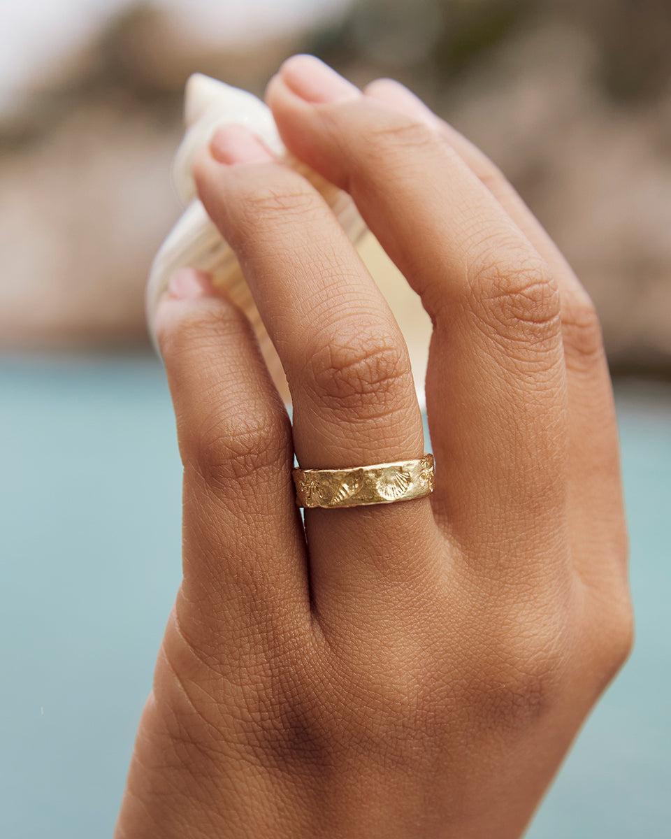 Gold Nucella ring with delicate coral and seashell engravings, worn on a finger, against a blue water background.