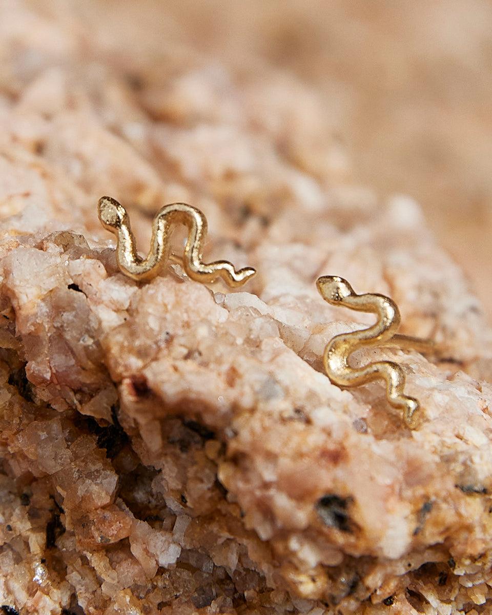 Petites puces d'oreille serpent dorées à la finition texturée, présentées sur une surface en pierre naturelle rugueuse.