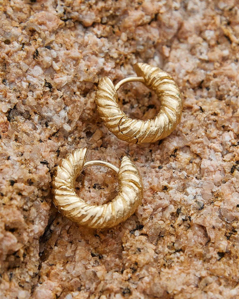 Pair of gold Zeta hoop earrings with twisted, rope-like texture on a textured rock surface.