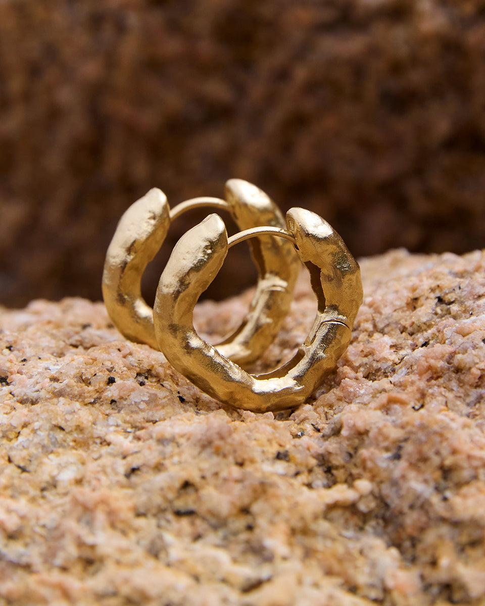 Pair of Adri gold geometric irregular hoop earrings with a textured finish, resting on a natural stone surface.