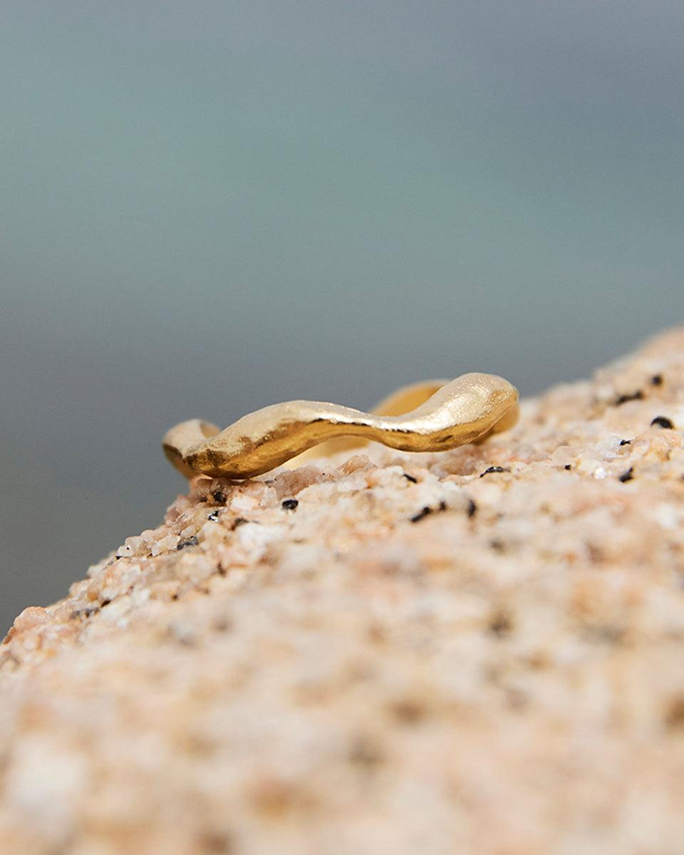 Thin, twisted gold Satis Ring with an undulating, wave-like design resting on a sandy rock.