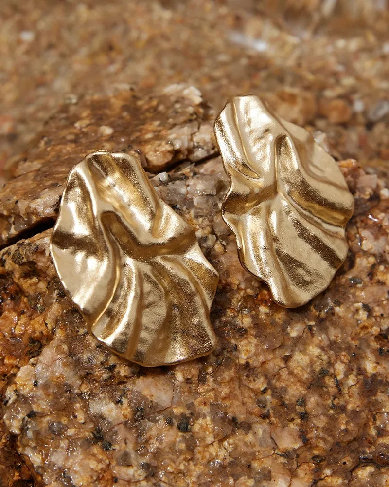 Pair of gold Batla earrings with a soft petal, fabric-like texture, resting on brown rocks.