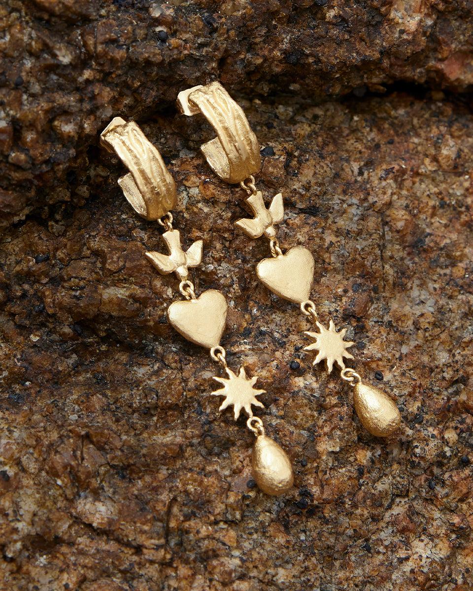 Gold Amara pendant earrings with dove, heart, sun, and drop charms on a textured rock background.