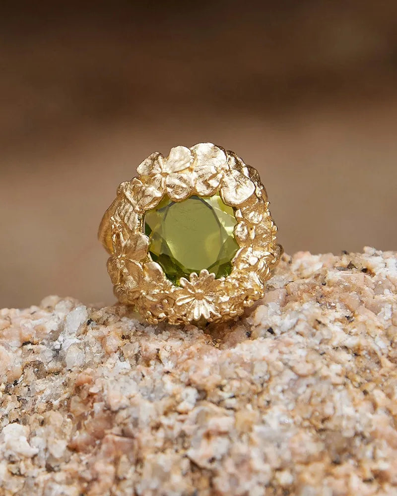 Gold Hespé chunky ring with a striking green stone surrounded by a bed of flowers, resting on a rock.