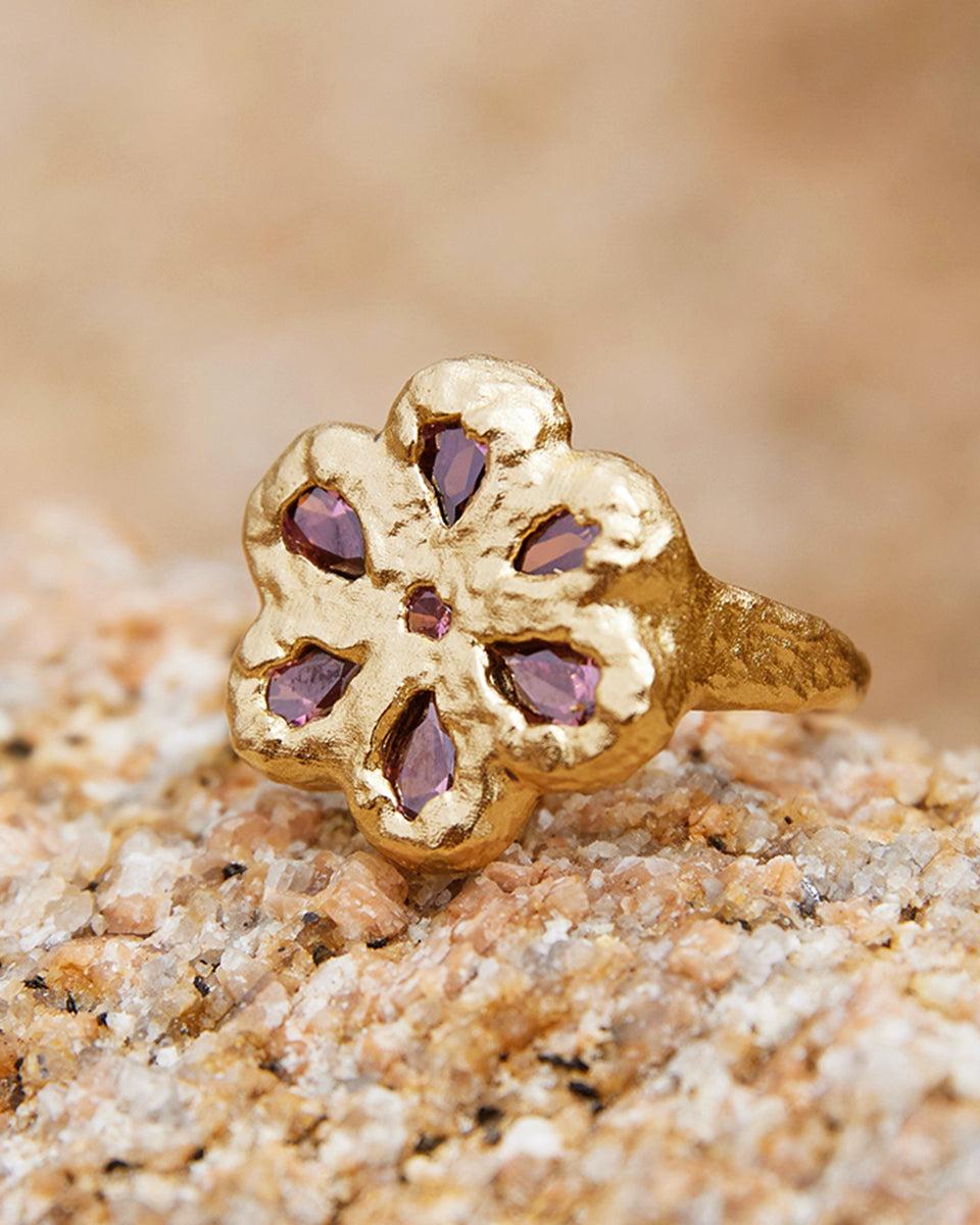 Rita Ring: Gold flower ring with five deep rosewood stones, resting on a textured rock.