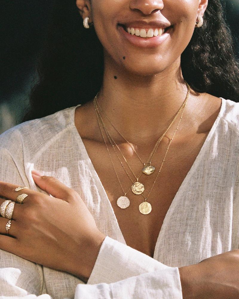 Gold Rafael moon pendant necklace with white stones, layered with two coin necklaces, worn by a smiling woman.
