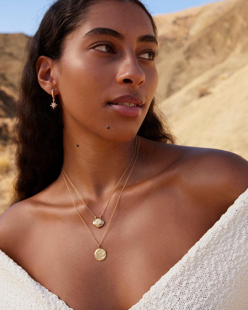 Woman wearing a gold Rafael moon pendant necklace with white stones, layered with a coin pendant, and a star earring.