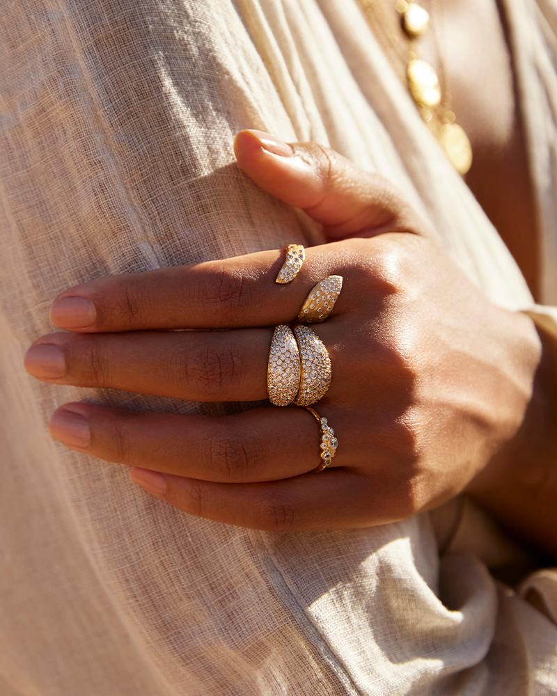 Breiter, gewölbter, goldfarbener Ring mit funkelnden, im Pavé gefassten, weißen Steinen, an der Hand einer Frau.