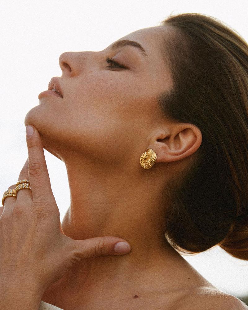 Gold Claudia chunky twist knot earrings with a ridged, domed, wave-like design on a model.