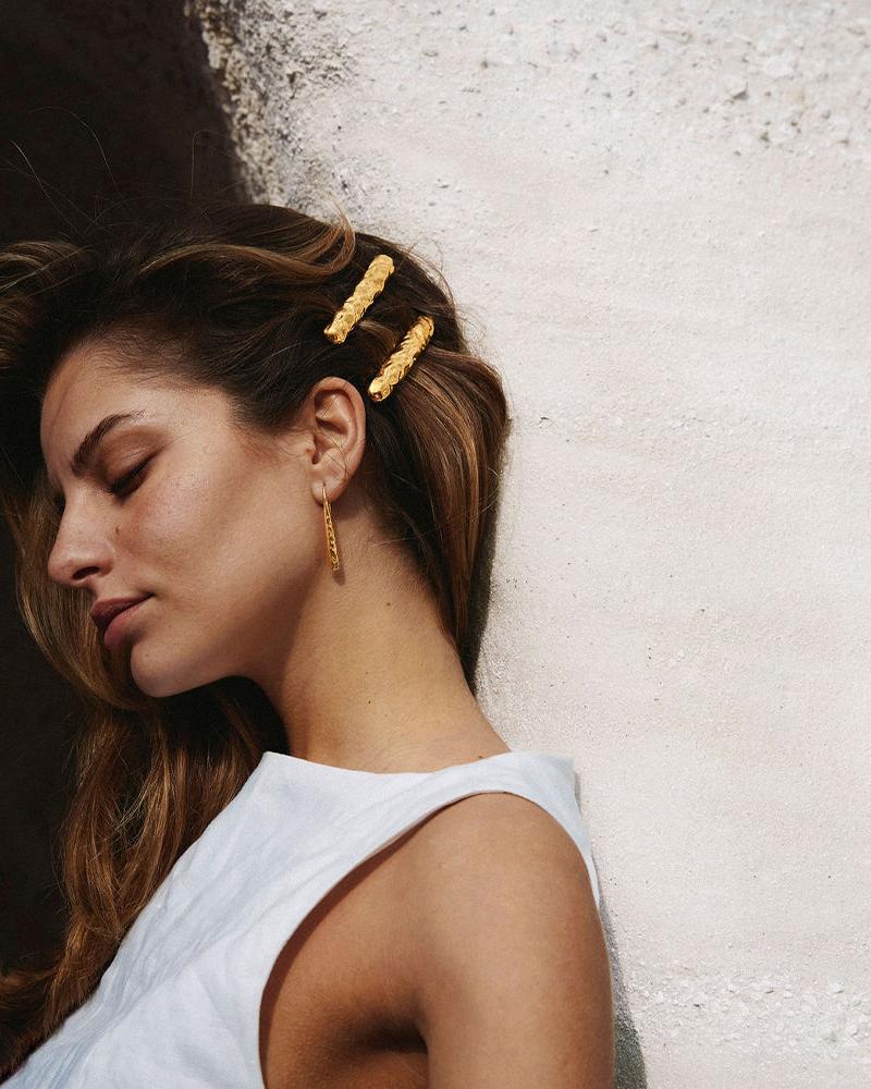 Two gold Nina hair clips with a hammered, organic texture worn in brown hair against a textured white wall.