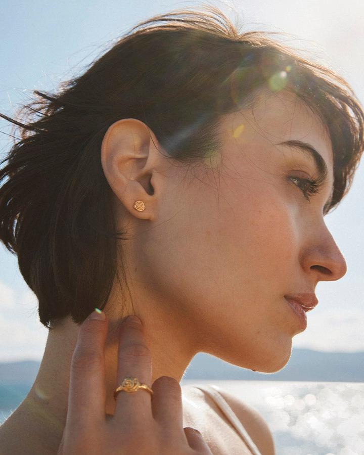 Model wearing small gold rose stud earrings with textured petals against a sunny coastal background.