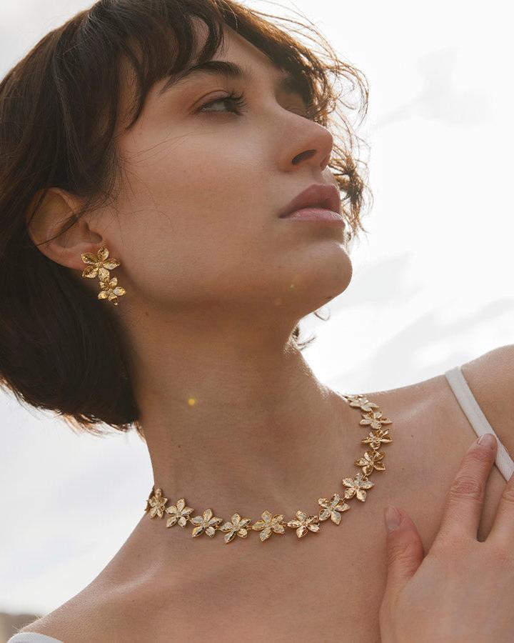 Thalla floral choker and matching earrings with hammered five-petal flowers in gold, worn by a woman.