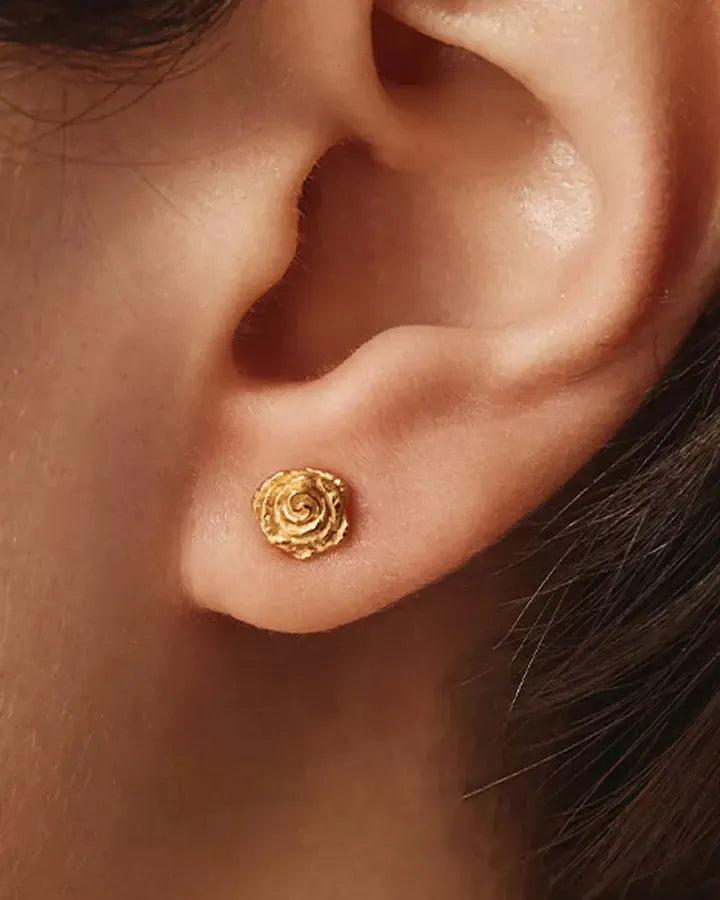 Petites puces d'oreille dorées en forme de roses miniatures aux pétales complexes et texturés, portées sur un lobe d'oreille.