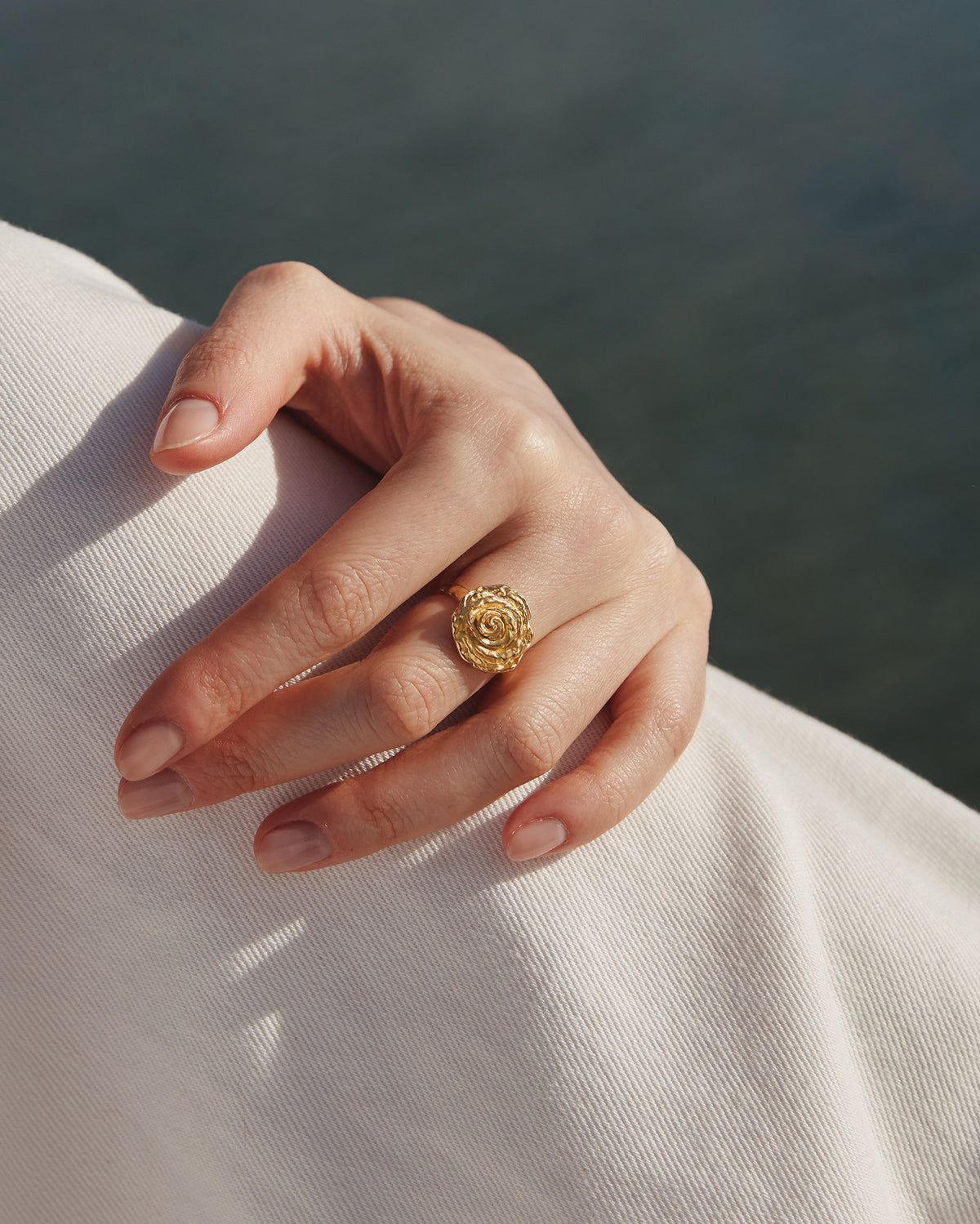 Gold statement ring featuring a sculpted rose with voluminous, hammered petals worn on a hand against white fabric.