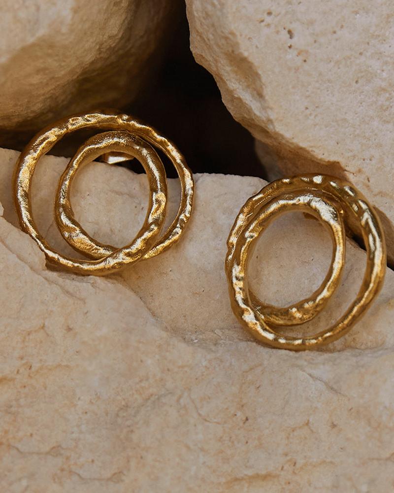Textured gold Orecchi earrings with a twisted, looping crossover design, resting on light-colored rocks.