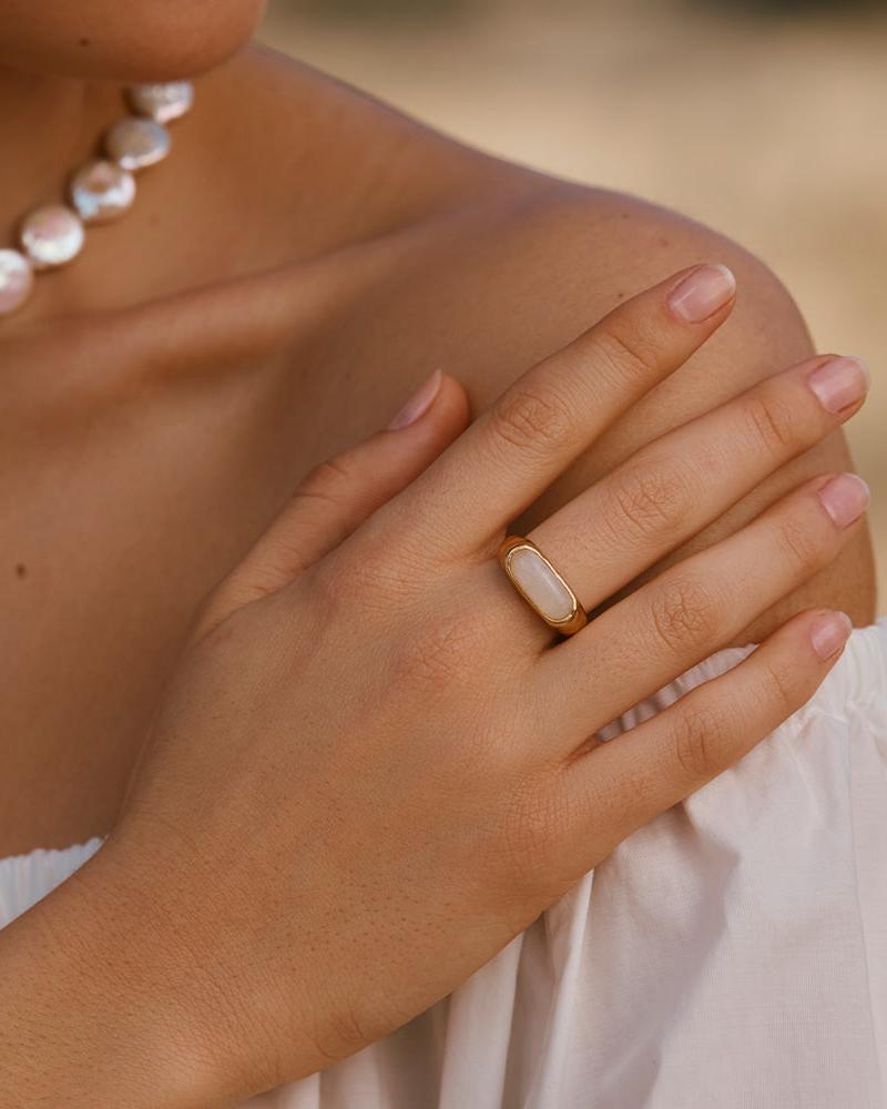 Gold Losna ring with an oval iridescent moonstone on a hand, worn with a pearl necklace.