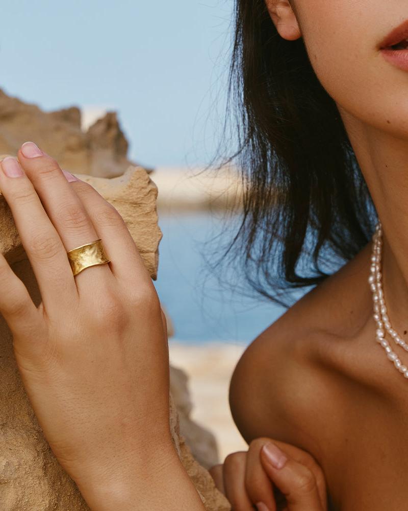 Gold Lucca ring, wide and concave with a hammered finish, worn on a hand against a coastal backdrop.