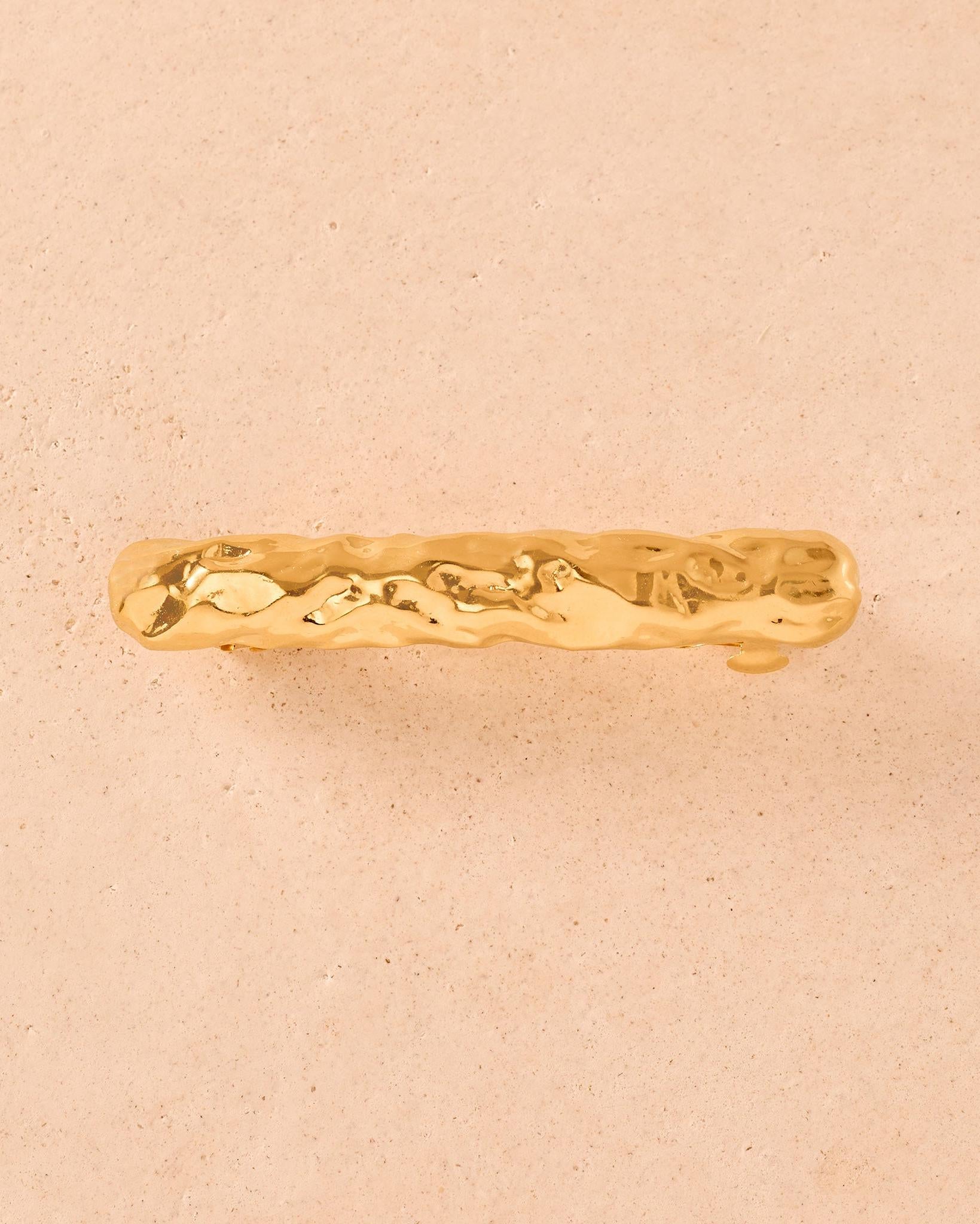 Gold Nina hair barrette with a hammered, organic metal texture on a light beige background.