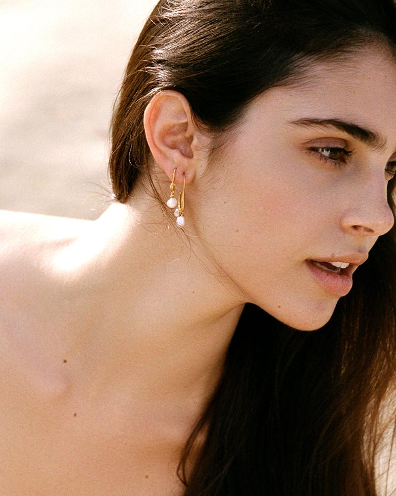 Gold Phaedra earrings with twisted open hoops and dangling freshwater pearl pendants on a woman's ear.