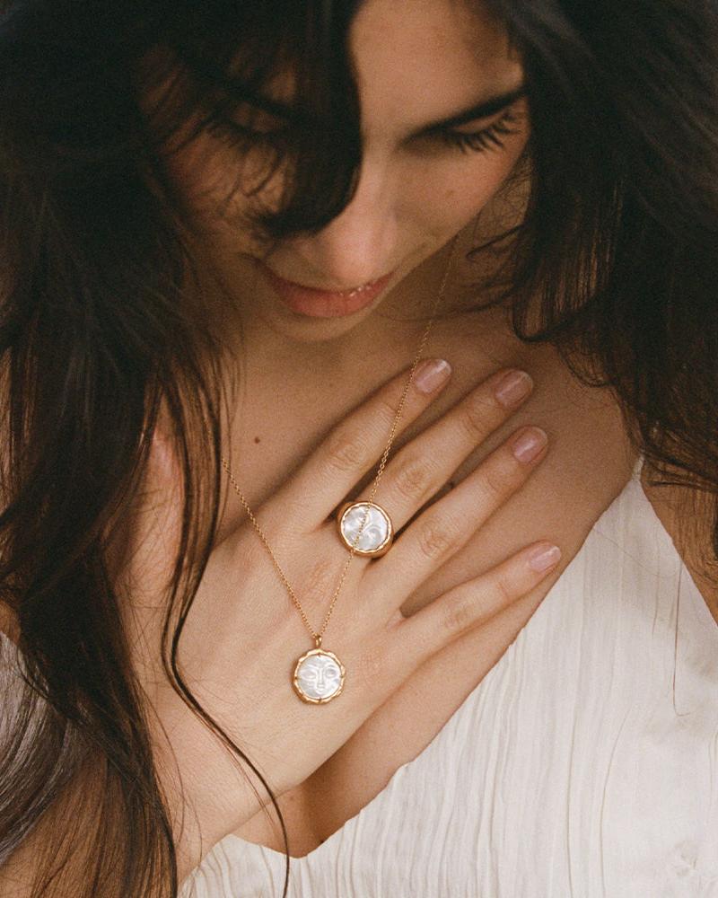Hëna necklace with two round, carved mother-of-pearl moonface pendants in golden settings on a woman's chest.
