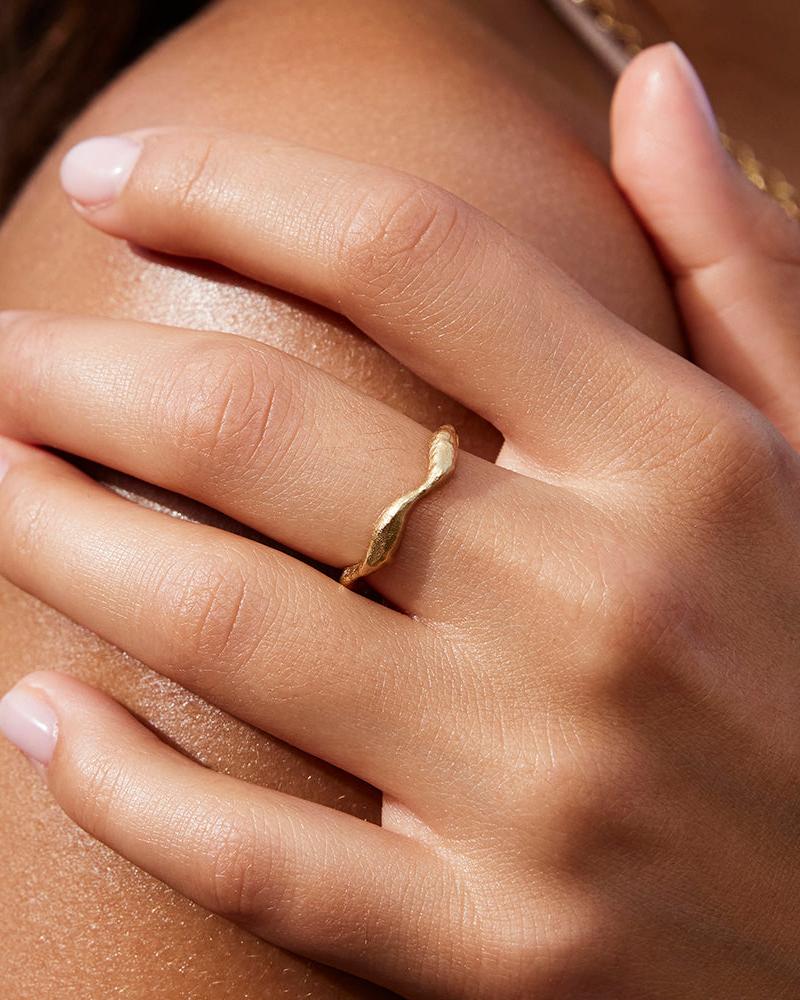 Dünner, gedrehter, goldfarbener Satis Ring mit wellenförmigem Design, an einer Hand.