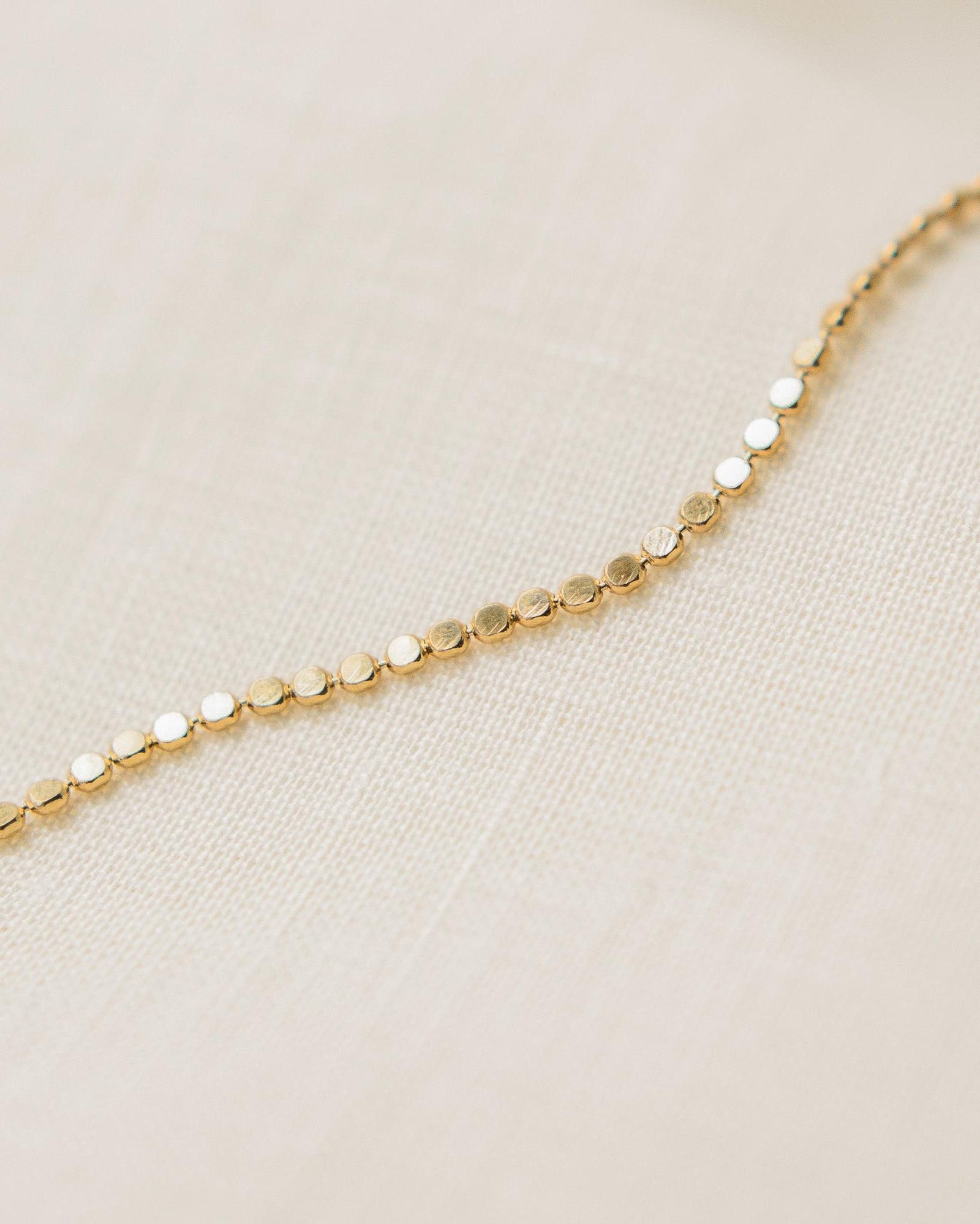 Delicate gold Anatole Bracelet with small, flat circular links on a light fabric background.