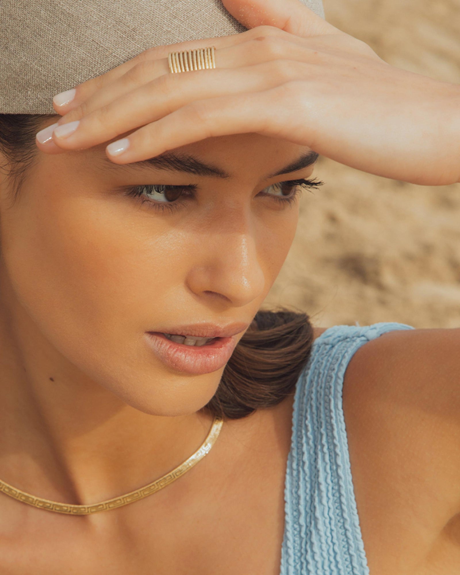 Close-up of a woman wearing a gold Anilla Large Earring, a gold necklace, and a beige hat.