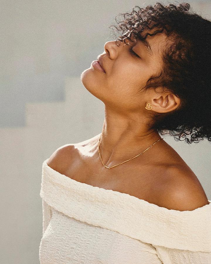 Woman wearing a Liora gold open-heart stud earring and a delicate gold necklace, eyes closed.