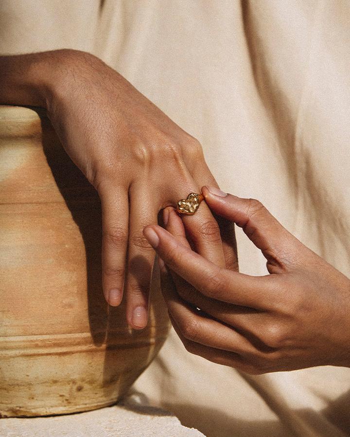 Gold Livia Ring with a chunky, hammered heart-shaped signet on a hand, next to an earthenware pot.