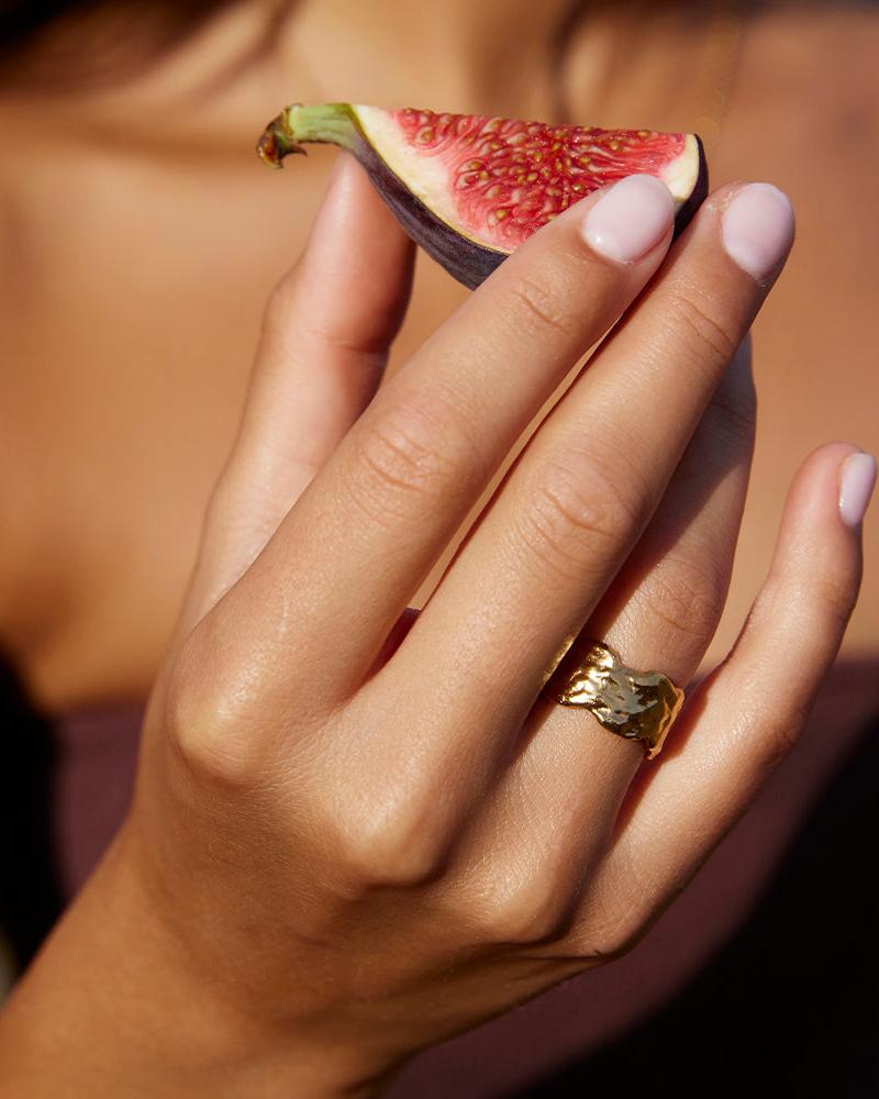 Anello a fascia larga Alphée dorato dalle linee organiche fluide e dalla texture martellata, su una mano che tiene un fico.