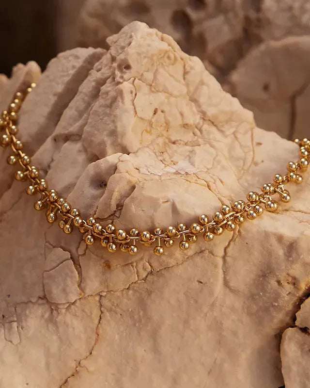 Achille gold-toned bracelet with small, round beads linked in a delicate chain, resting on a light-colored rock.