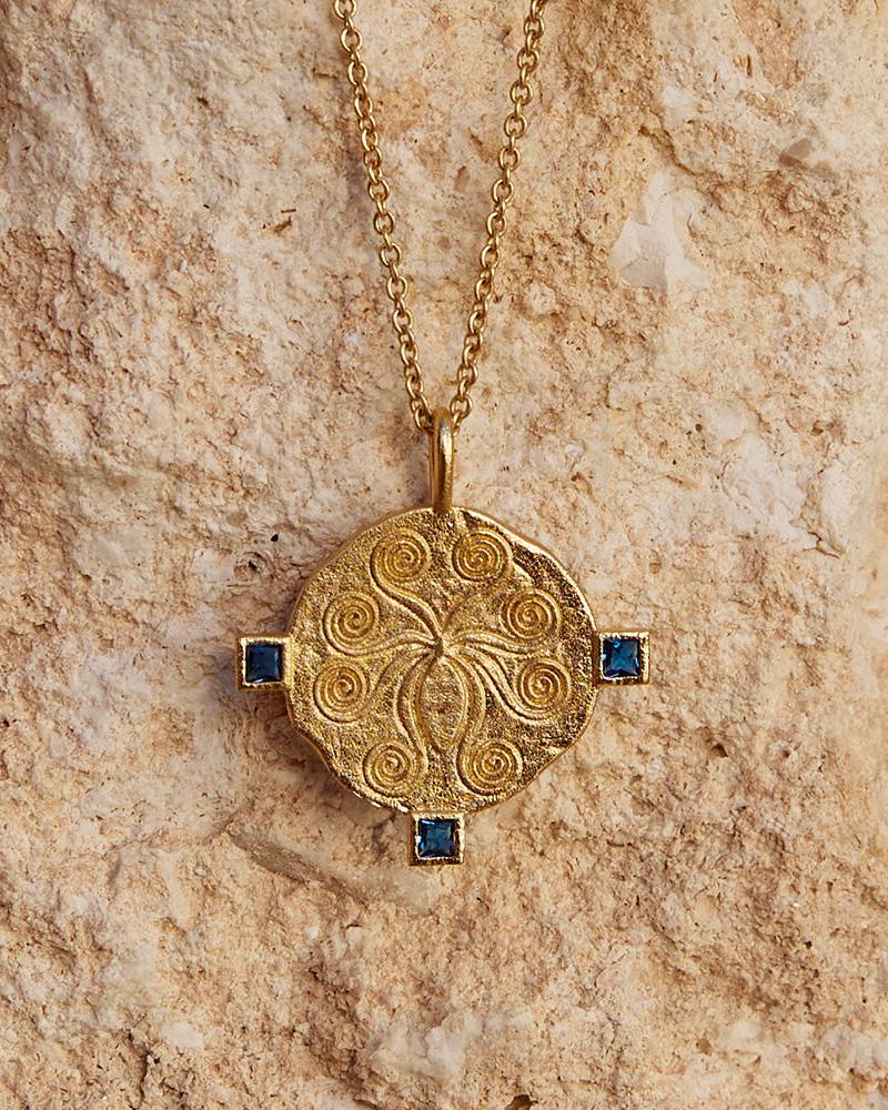 Gold Oxta necklace with an octopus design and four square blue gemstones on a textured background.