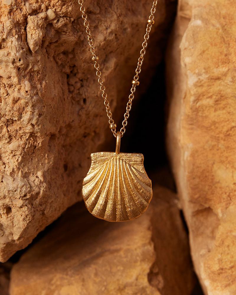 Milos Necklace: Gold seashell pendant on a delicate beaded chain, nestled between textured rocks.